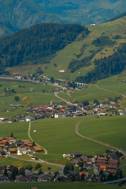 A scenic rural landscape featuring a narrow, paved country road lined with a wooden fence on both sides, leading towards a wooden house situated on a grassy hillside. The house has a dark brown exteri