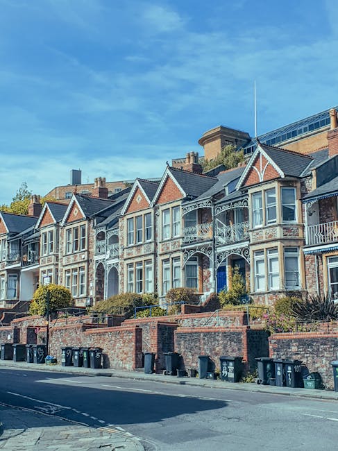 A row of Victorian-style terraced houses with intricate white ironwork balconies and decorative trim, situated along Finchley Road in Swiss Cottage. The houses are built with brick and painted wood, featuring bay windows and gabled roofs, set on a slightly elevated brick front garden with small shrubs and trees. The pavement below hosts black wheelie bins lined up along the curb, and a street with parked cars is visible in the foreground. The sky is clear with a few wispy clouds. This scene may be part of an illustrative context for home relocation or furniture transport services by Swiss Cottage Man and Van, highlighting the residential environment where packing and moving activities, such as loading furniture and packed boxes, could take place.